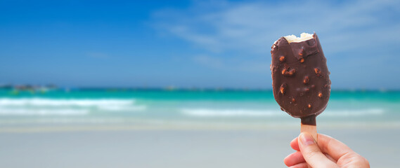 Obraz premium Chocolate covered ice cream bar in the summer at the beach, during the holidays. Hand holds delicious chocolate and nuts almond ice cream bar on a stick.
