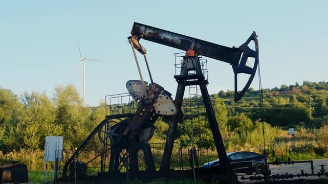 Rusty pumpjack rises before hillside while parked car stands nearby. Beam pivots with horsehead dips and lifts during pumping stroke