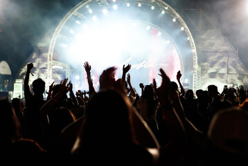 Silhouettes of hand raised of fans in crowd at concert in front of stage with bright spotlights.The back view of people enjoy live music at the band's performance. Outdoor concert.