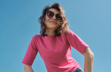 Beautiful carefree young woman in sunglasses and pink t-shirt smiling on blue sky background low angle shot.