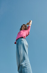 Beautiful carefree young woman with curly hair in pink t-shirt raising her hands up on blue sky background low angle shot.