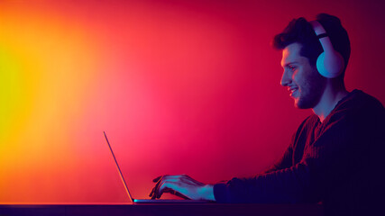 Man using laptop with headphones in neon digital environment. Concept of focused workflow, online meetings, remote communication and technology powered concentration.
