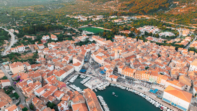 Aerial view of terracotta rooftops cascading towards the turquoise harbor where boats bob gently, framed by lush green hills, Cres, Primorsko-goranska zupanija, Croatia.