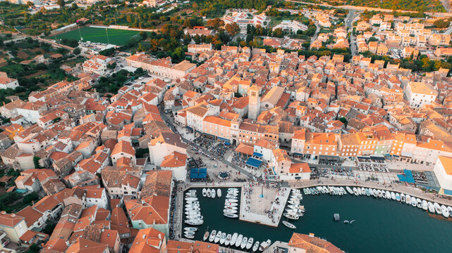 Aerial view of sun-kissed terracotta roofs cascade towards a bustling harbor filled with white boats, framed by historic buildings, Cres, Primorsko-goranska zupanija, Croatia.