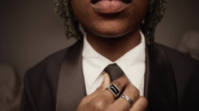 Tilt up portrait shot of young African American man wearing formal suit and stylish eyeglasses adjusting black tie while rapping at camera during studio music video shoot, slow motion