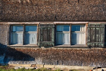 Facade of a traditional building with shingles, giving the building a rustic appearance. There are two windows with white curtains and weathered, dark-painted shutters.