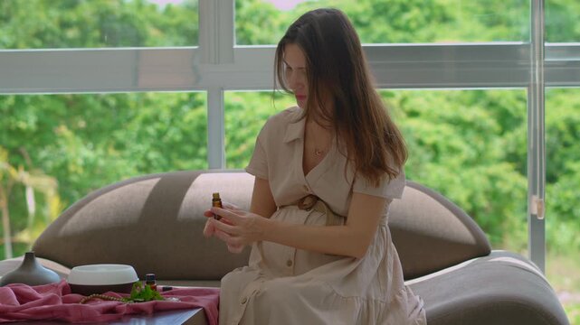 Pregnant woman over 40 sitting on a couch by a sunlit window enjoying scent and relaxation from a working electric aroma diffuser. Wellness, pregnancy care and relaxation lifestyle concept. Slowmotion