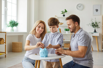 Fototapeta premium Happy young family parents and child sitting on floor in living room and putting coins in blue piggy bank standing on table at home. Smiling mother, father and son planning family budget together.