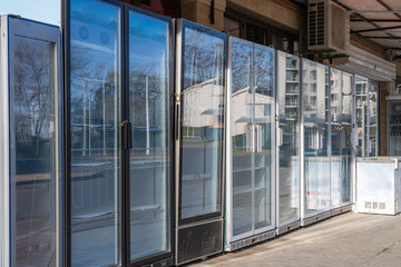 a row of empty commercial glass-door refrigerators and freezers standing on a paved sidewalk outside a building, with reflections of the surroundings visible on the glass.