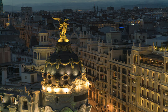 Aerial view of the Metropolis Building illuminated against the night sky, its golden statue gleaming, amidst a dense urban landscape, Madrid, Community of Madrid, Spain.
