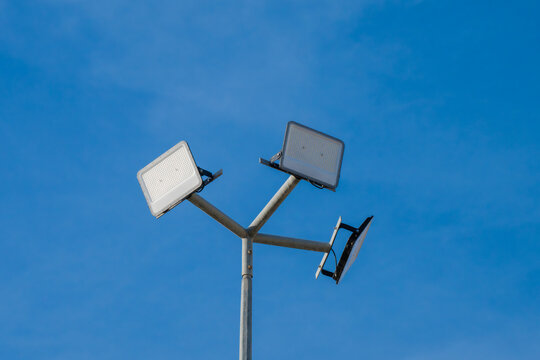 a metal light pole supporting three rectangular LED floodlights arranged on arms against a clear blue sky.