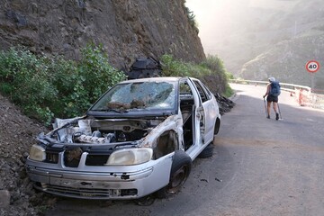 Hermigua, La Gomera, Spain : A car destroyed by a falling stones stands on mountain asphalt road. Silhouettes of hiking people on road. Canary Islands © Iwona