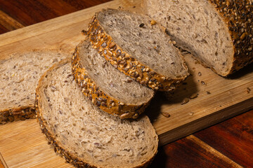 A close-up view of a loaf of seeded whole grain bread with several slices cut, resting on a wooden cutting board with scattered crumbs.