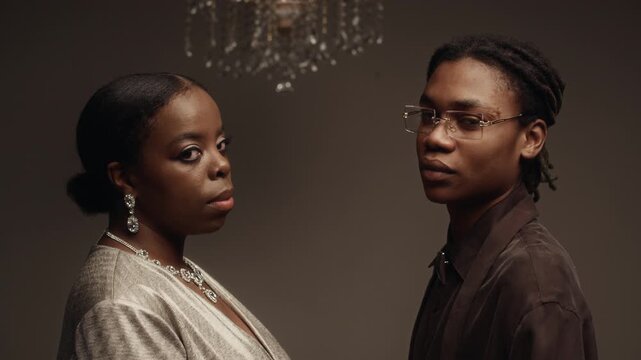Profile view of Black woman wearing rhinestone necklace with matching earrings looking at Black man in stylish glasses then turning to camera while posing against mocha colored studio wall