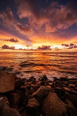 Sunset over ocean with waves hitting rocky beach under dramatic orange clouds. Golden hour seascape with sea, shoreline, and sky, ideal for travel, nature, background, and coastal themes.