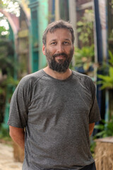 A man with a beard and a gray shirt is standing in front of a building. He looks serious and is looking up at something