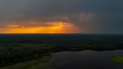 Aerial view of a vast forest landscape with a serene lake reflecting the dramatic sunset. Soft orange hues contrast with dark clouds, creating a tranquil yet captivating atmosphere.