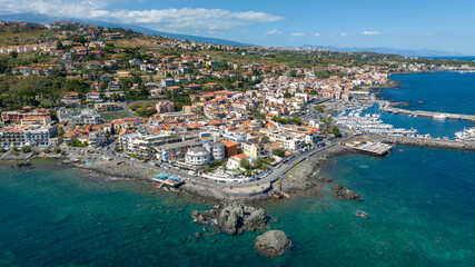 Fototapeta premium Aerial view of the town of Aci Trezza overlooking the Mediterranean Sea, in the province of Catania, Sicily, Italy. In the background is the marina of the city. It's a sunny summer morning.