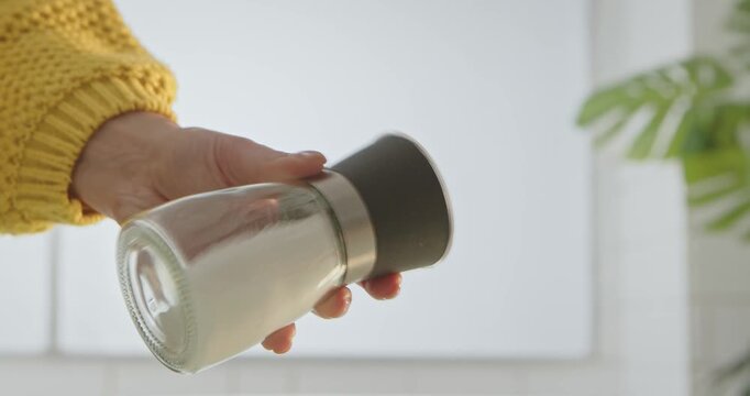 Close up of a hand in a yellow sweater shaking a clear glass salt mill with a metal lid indoors, preparing to season food.