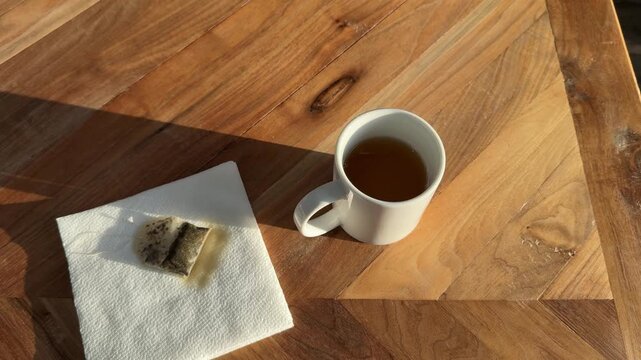4K video of a hand removing a tea bag from a mug and placing it on a napkin in warm natural morning light on a wooden table, simple tea routine and cleanup concept.