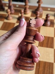 A hand holding a wooden chess piece (king), with a classic French manicure on the nails. Blurred image, business woman hand holding chess price on chess board. Competition success and strategy game