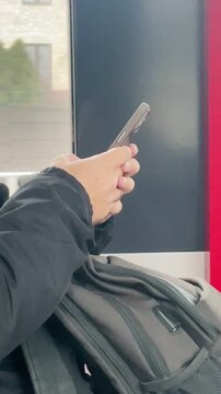 hands texting on phone above backpack beside train window, scrolling through messages with landscape blur outside. Vertical shot taken on mobile device