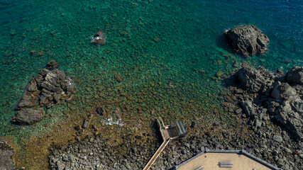 Aerial view of a rocky coast with a crystalline sea. The colors of the water fade from blue to emerald green. There are rocks that emerge near the shore.