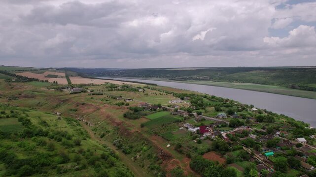 Aerial River Valley Village Under Overcast Sky, Winding River Bordering Green Banks, Scattered Rural Houses And Farm Plots, Unpaved Roads Cutting Through Pastures, Moody Cloudscape