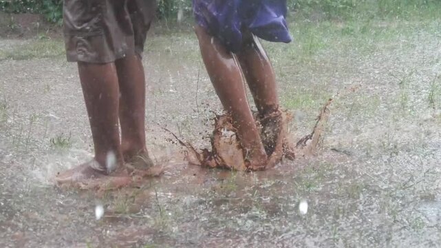 The feet of two boys playing and jumping in a muddy puddle during the rain. The splashing mud and water create a cheerful atmosphere.