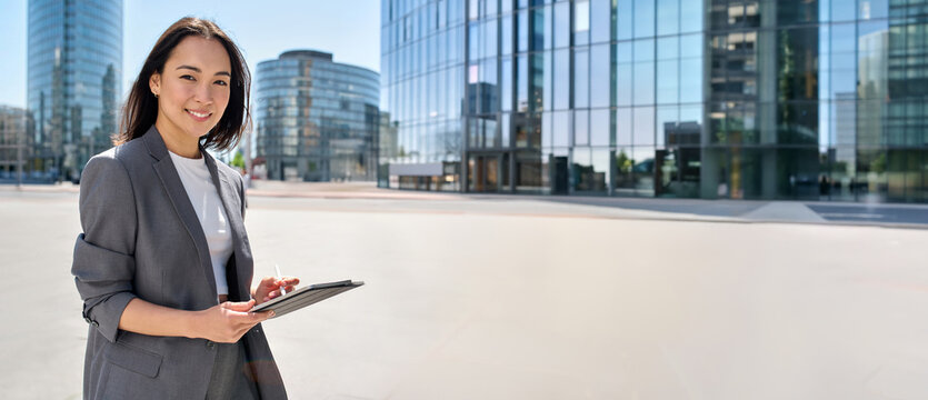 Young happy Asian business woman wearing suit holding digital tablet standing in big city on busy street, using smart business software tech for online work on pad computer. Banner, copy space