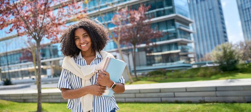 Happy pretty female African American girl student standing outside university holding notebooks looking at camera outdoors, portrait. Applying university, admission and scholarship program. Banner