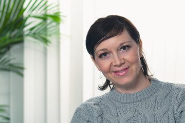 A woman with brown hair and a gray sweater is smiling. She is sitting in front of a plant
