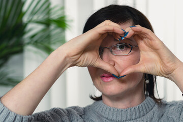 A woman wearing glasses and blue nail polish is making a heart shape with her hands. Concept of warmth and affection, as the woman's gesture suggests love and care