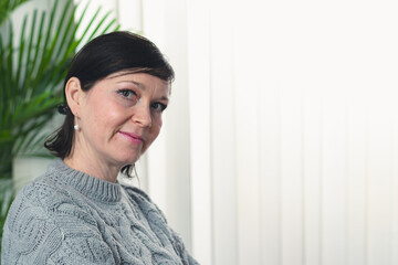 A woman is sitting in a chair with a plant behind her. She is wearing a gray sweater and has her hair pulled back