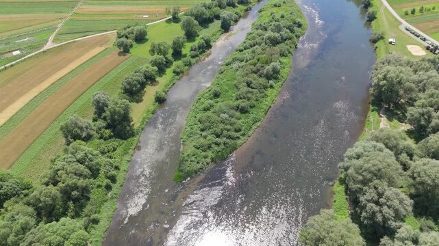 Aerial view of a braided river flowing between cultivated fields with visible current movement and green islands forming natural patterns across a rural summer landscape