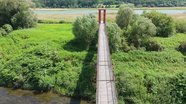 Top down aerial view along a narrow suspension bridge spanning a shallow river with steady water movement and dense vegetation on both sides under clear daylight