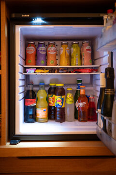 A hotel minibar stocked with assorted bottled drinks including Coca-Cola, Fanta, Lipton iced tea, Schweppes, Peroni, and Aperol Spritz, neatly arranged inside a small illuminated refrigerator.