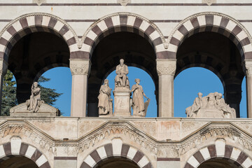 Obraz premium Marble statues of allegorical figures and seated man displayed between striped arches on façade of Monumental Cemetery arcade in Milan, Italy.