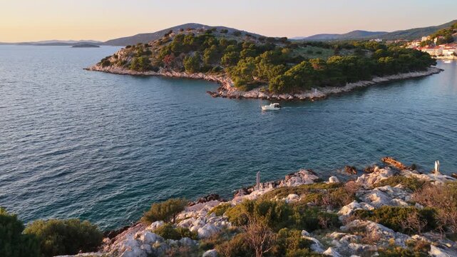 Aerial drone view of the rugged coastline near Tribunj, Croatia , featuring crystal clear waters, rocky shores, low Mediterranean vegetation, and a fishing boat passing between the peninsula