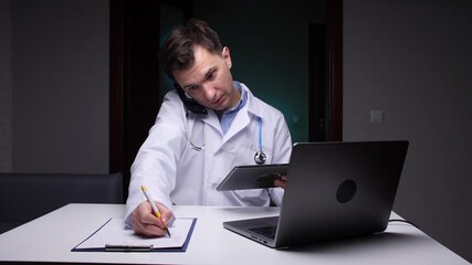 Medical professional wearing a lab coat and a stethoscope concentrating on work tasks at a desk, writing notes, answering a phone call, and using a laptop and tablet in a dark office setting - Powered by Adobe