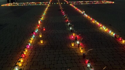 Memorial installation of colorful burning candles arranged in a large christian cross on a cobblestone square at night, honoring the memory of victims and celebrating faith and hope