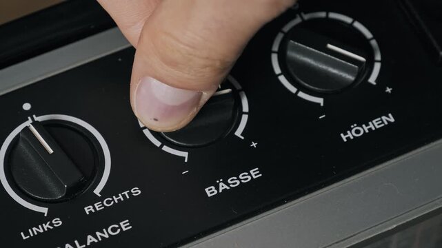 Close-up of a male hand adjusting a black rotary bass knob on a vintage German audio player. White markings change low frequency sound levels on the stereo system.