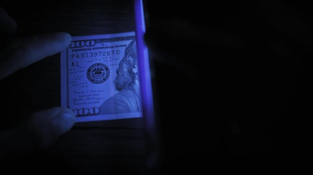 Overhead view of a hand holding a one hundred dollar bill under an ultraviolet lamp for verification. The glowing security thread confirms authenticity against a dark wooden surface.