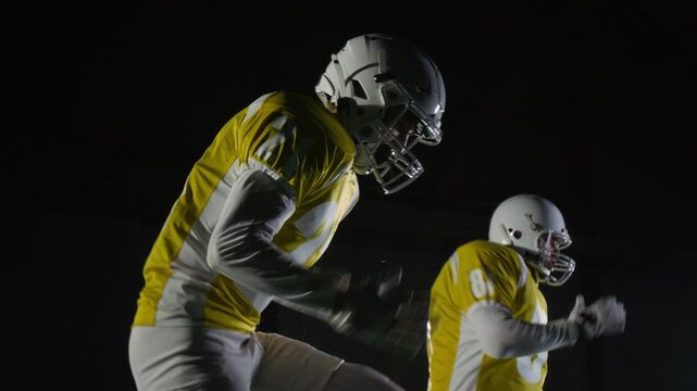 Medium side shot of team of American football players, wearing protective padded gear, helmets and jerseys, jogging on spot to warm up while getting ready for training session