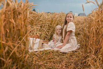 Two young girls sit in a golden wheat field. The older girl has long brown hair and a light dress. The younger girl has short blonde hair and a similar dress.
