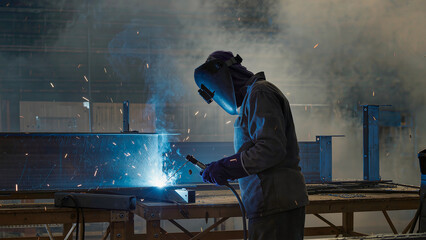 A worker in a full protective welding helmet and suit performing arc welding on large steel beams in a factory. Bright blue light and golden sparks illuminate the smoky industrial environment. Concept