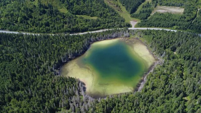 Summer aerial video moving away from a small turquoise lake near the road passing at the foot of the mountain in Val-d'Irene Regional Park, a ski resort in winter. Sainte-Irene, Quebec, Canada, 2025.