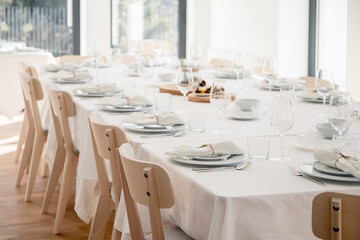 A bright elegant dining hall is prepared for a festive gathering, with a long white table neatly set with plates, glasses and linen napkins, ready to welcome guests for a celebratory meal.