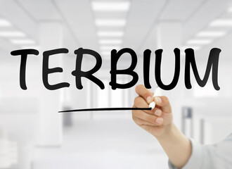 A conceptual image showing the word terbium handwritten in bold black letters on transparent glass, with a blurred modern white office interior in the background, symbolizing this rare earth element&rsquo;s