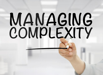 A conceptual business image showing the phrase managing complexity handwritten in bold black letters on transparent glass, with a blurred modern white office interior in the background. The clean corp